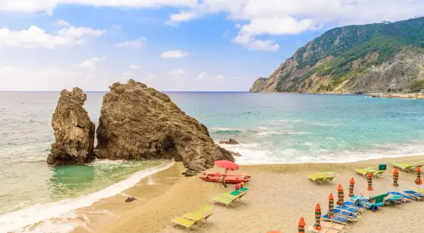 Spiaggia sabbia Liguria con scogli imponenti, mare azzurro, lettini e ombrelloni pronti per accogliere i bagnanti.