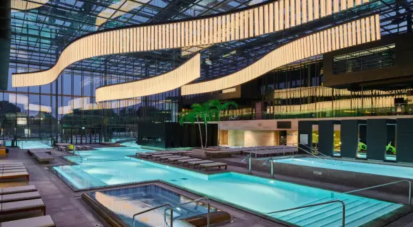 Interno moderno delle Merano Terme con piscine luminose, lettini e palme sotto un soffitto vetrato con illuminazione scenografica.