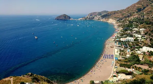 Vista aerea di una baia con spiaggia affollata, ombrelloni colorati e mare blu intenso. Un promontorio roccioso e un isolotto punteggiano l'orizzonte. Potrebbe essere una delle 5 spiagge più belle di Ischia.