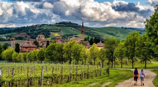 Due escursioniste passeggiano tra vigneti rigogliosi e dolci colline modenesi, con un borgo antico sullo sfondo.