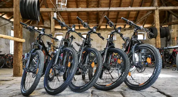 Bici in montagna pronte per l'avventura. Diverse mountain bike nere con dettagli arancioni, parcheggiate in un fienile con travi in legno.