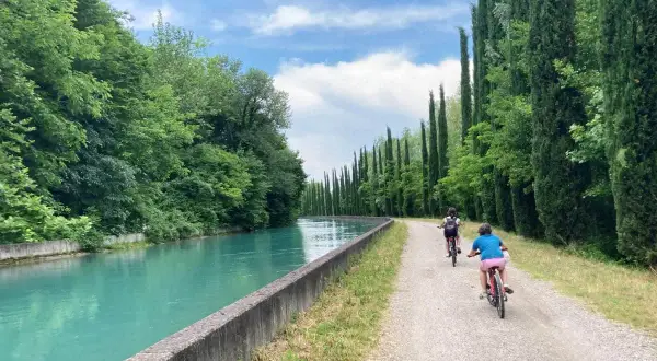 Bambini in bicicletta lungo la ciclabile Mincio, con un canale turchese e filari di cipressi.
