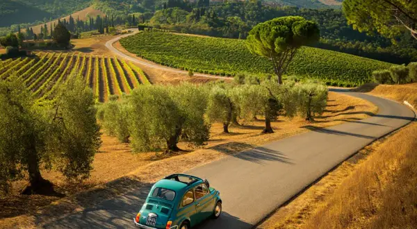 Un'iconica Fiat 500 azzurra percorre una strada tortuosa tra vigneti e ulivi nei **colli bolognesi**, con borghi arroccati all'orizzonte.