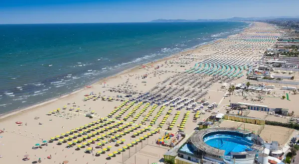 Vasta spiaggia della **costiera romagnola** con file ordinate di ombrelloni gialli e azzurri, mare calmo e un complesso con piscina.