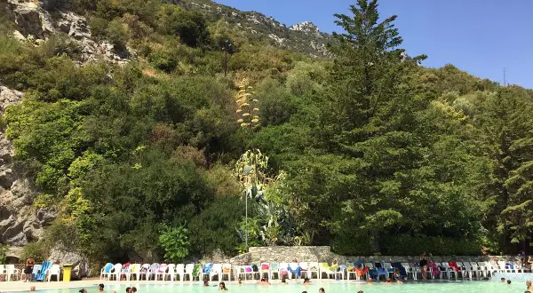 Piscina con persone che nuotano, circondata da vegetazione lussureggiante e rocce, un luogo che sembra uscito dalle grotte delle ninfe.