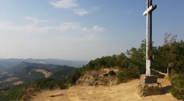 Croce in cima a un crinale roccioso, con vista panoramica sui monti dell'Emilia Romagna e un cielo azzurro con nuvole bianche.