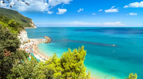 Spiaggia affollata con ombrelloni colorati e gente che nuota nelle acque cristalline. Una scogliera bianca domina la costa, incorniciata dal verde della vegetazione. Una perfetta località mare Marche.