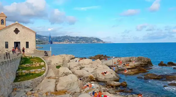 La chiesa bianca domina la costa rocciosa di Bordighera, con bagnanti che si godono il sole. Un luogo incantevole per scoprire cosa vedere a Bordighera.
