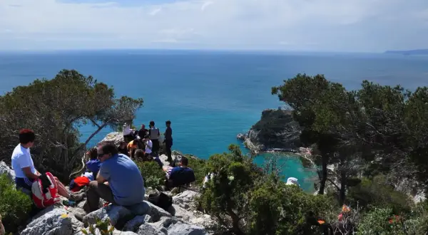 Gruppo di persone in pausa durante un trekking a Finale Ligure, con vista sul mare azzurro e scogliere.