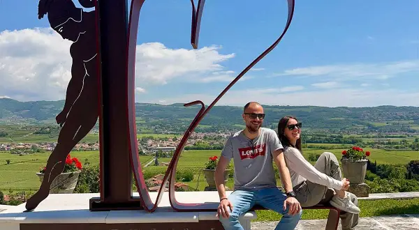 Co zobaczyć w Valpolicella: para siedzi na ławce przed rzeźbą serca i postacią, z widokiem na winnice i wzgórza.