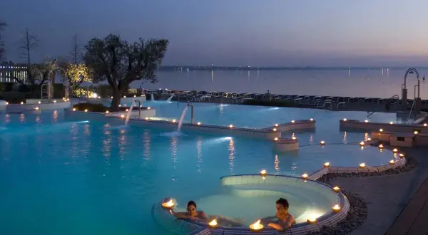 Coppia rilassata in una piscina termale illuminata da candele, con cascate e vista lago al tramonto. Un'oasi di pace alle terme a Brescia.