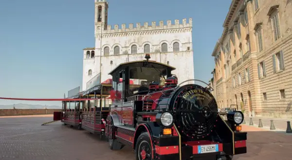 Il trenino turistico di Gubbio, rosso e nero, attende i passeggeri in una piazza storica con palazzi antichi.