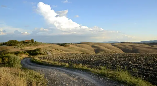 Strada sterrata serpeggia tra le dolci colline senesi, con un casale in lontananza sotto un cielo azzurro e nuvoloso.