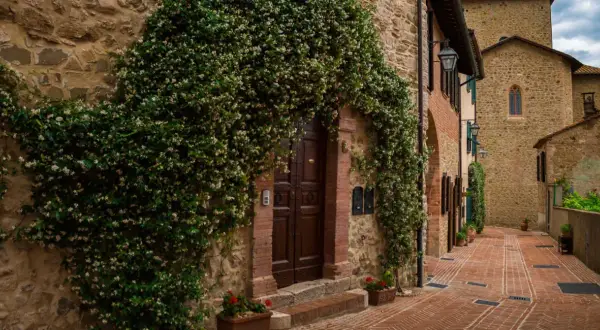 Un antico borgo in Umbria, con un portone di legno scuro avvolto da rampicanti fioriti. La strada in mattoni rossi sale dolcemente tra case in pietra.