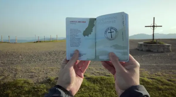 Un passaporto dell'Appennino Modenese, con la Croce Arcana di Fanano, tenuto in mano davanti a un panorama montano e una croce.