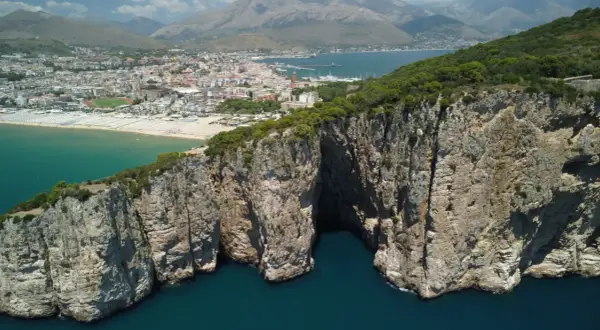La leggenda della Montagna Spaccata a Gaeta: imponenti scogliere rocciose si tuffano nel mare blu, con la città sullo sfondo.