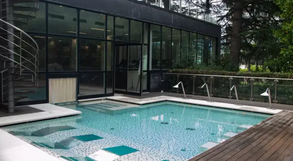 Piscina moderna con mosaico azzurro e bianco, scala a chiocciola in metallo, vetrate e alberi rigogliosi. Un'oasi di relax a Boario Terme.