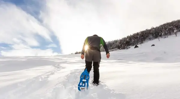 Escursionista con racchette da neve cammina su sentiero innevato, scoprendo posti belli in montagna Piemonte inverno. Sole splendente tra le nuvole.