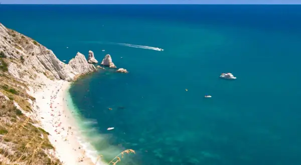 La spiaggia più bella del Conero, con sabbia bianca, mare cristallino e scogliere imponenti. Barche solcano le acque turchesi.