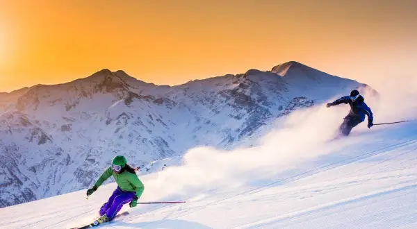 Due sciatrici sfrecciano su un pendio innevato al tramonto, con montagne maestose sullo sfondo. Un'emozione da provare per chi cerca dove sciare in Emilia Romagna.