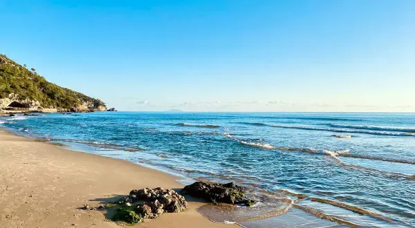 Spiaggia dorata e mare azzurro vicino a Roma. Le onde lambiscono rocce coperte di alghe, mentre una collina verde domina la costa.