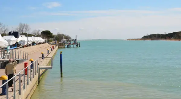 Un uomo guarda il mare calmo a Marano Lagunare, con barche coperte e un molo. La spiaggia lagunare è serena.