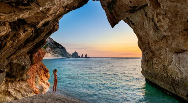 Vista da una grotta sulle spiagge della Riviera del Conero al tramonto. Una ragazza ammira il mare turchese e le rocce a picco.