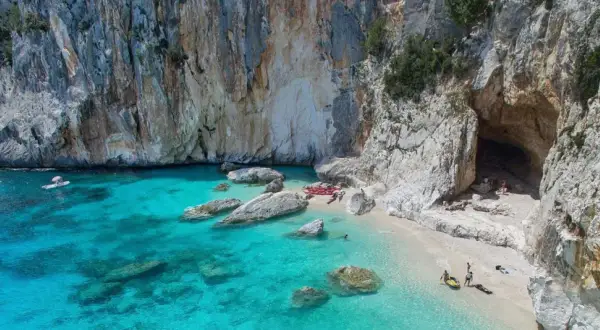 Spiaggia incantevole sulla costa di Baunei, con acque cristalline, rocce imponenti e una grotta marina. Kayak e persone godono del sole.