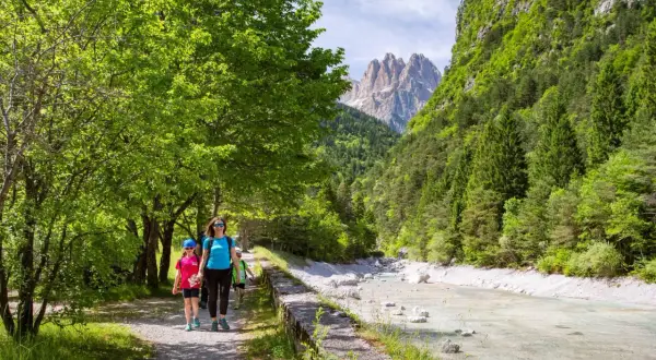 Famiglia in escursione ad Andalo, cammina lungo un sentiero con un fiume cristallino e montagne maestose sullo sfondo.