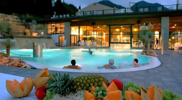 Piscina termale illuminata di notte, con persone che nuotano e un tavolo di frutta fresca in primo piano. Un'oasi di relax che ricorda le terme di Reggio Emilia.
