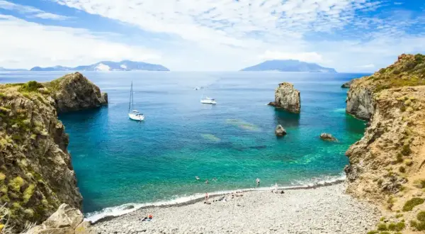 Spiaggia di ciottoli e acque cristalline, ideale per una vacanza alle Eolie. Barche a vela e isole all'orizzonte.