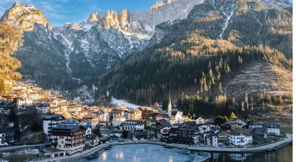 Villaggio alpino innevato ai piedi delle Dolomiti, ideale per escursioni invernali. Lago ghiacciato riflette le montagne.
