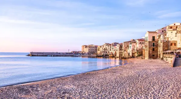 La spiaggia di Cefalù al tramonto, con le case colorate che si affacciano sul mare calmo e la sabbia dorata.