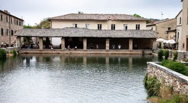 La piazza delle sorgenti termali di Bagni Vignone, con persone che si rilassano sotto un portico e il riflesso dell'acqua.