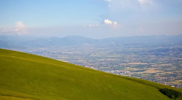 Prato verde scende dolcemente dal monte Subasio verso una valle punteggiata di case e montagne all'orizzonte.