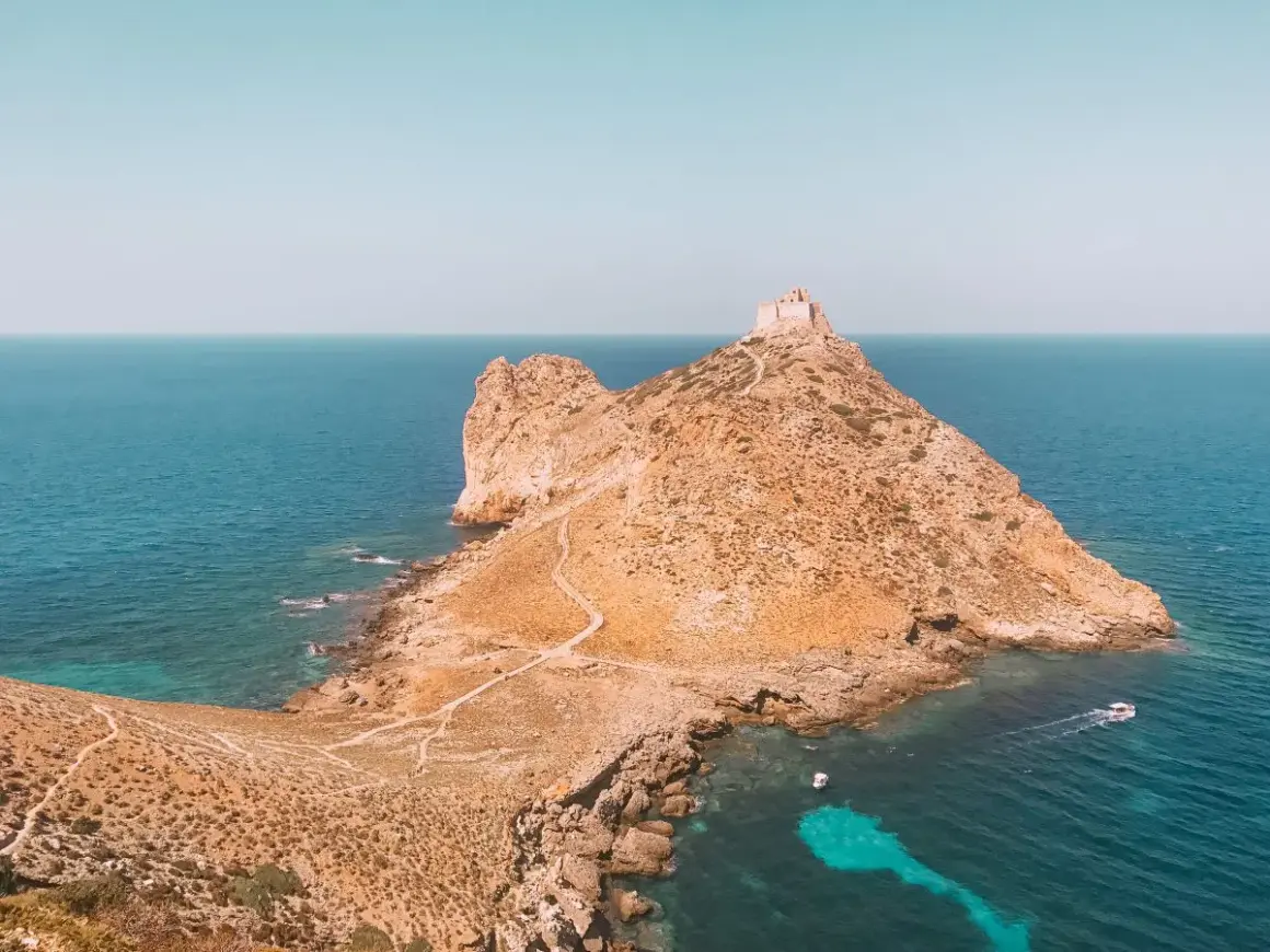 Rocciosa isola con un antico fortino in cima, circondata da acque turchesi. Un sentiero serpeggia verso la vetta, evocando le spiagge delle isole Egadi.