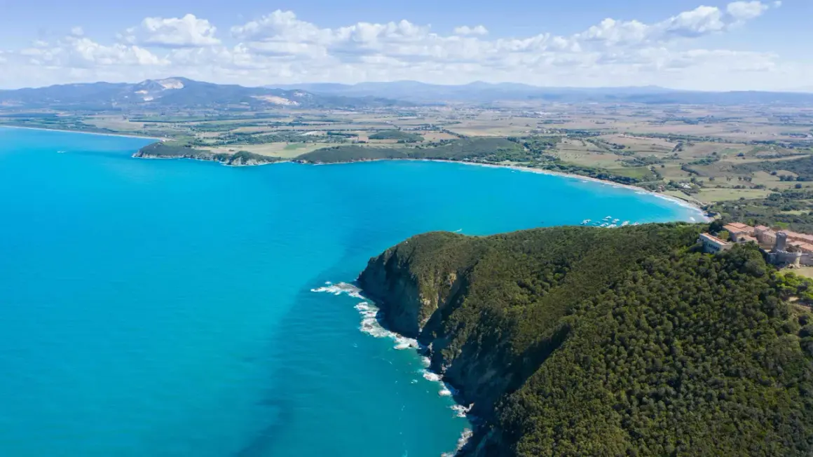 Vista aerea della costa degli Etruschi con mare azzurro, scogliere verdi e un borgo storico.