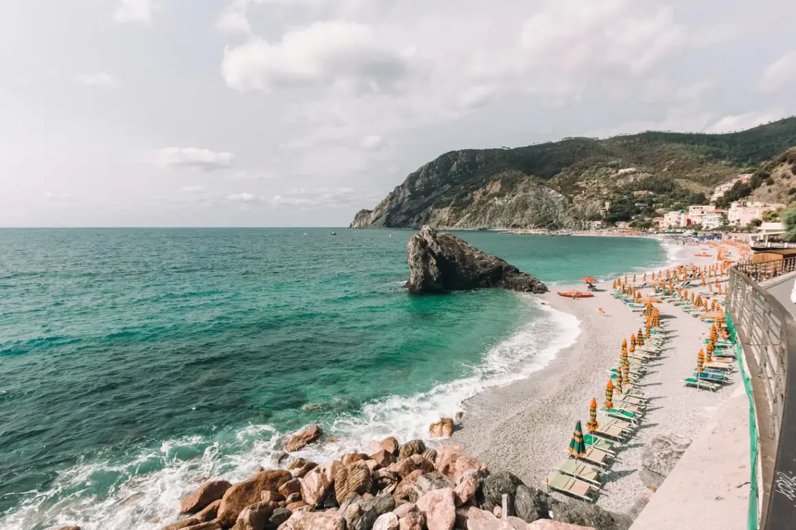 Spiagge libere Liguria: mare turchese, scogli imponenti e file di ombrelloni colorati su una spiaggia di ciottoli.