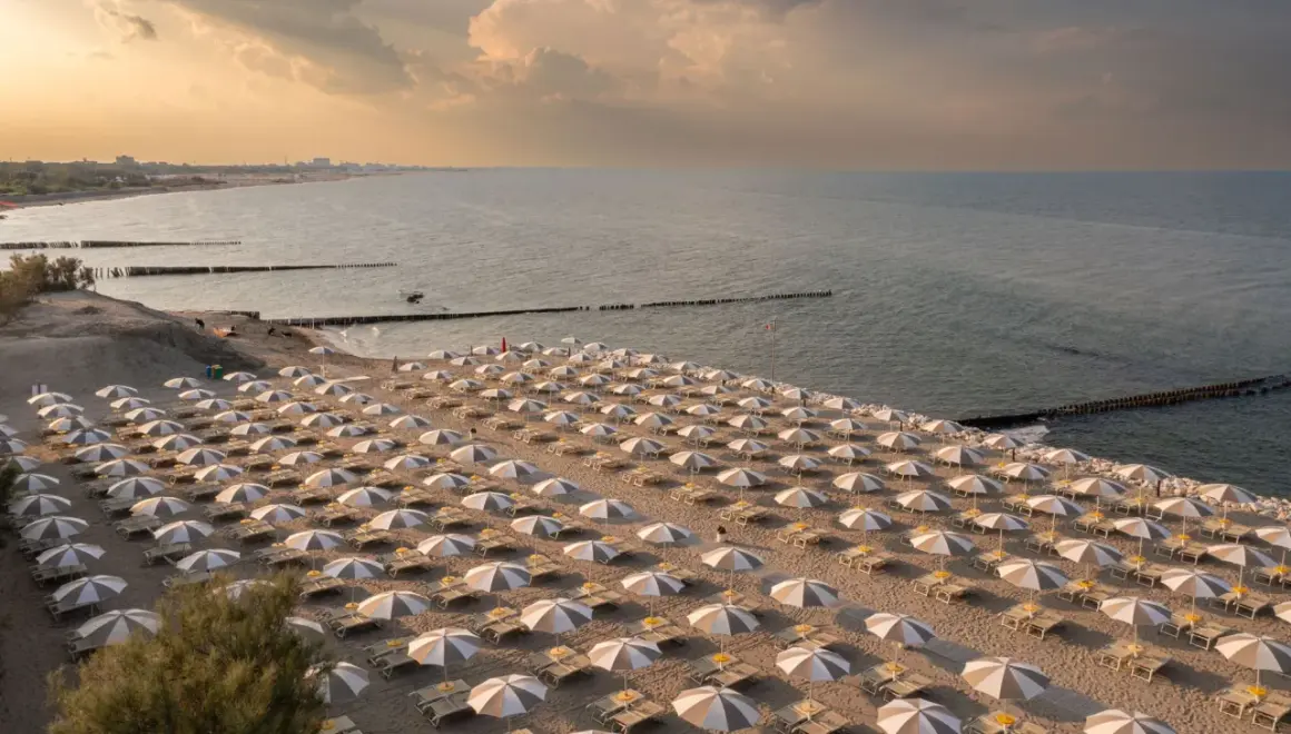 Filetto di sabbia dorata con file ordinate di ombrelloni e lettini, bagnato dal mare del Friuli Venezia Giulia al tramonto.