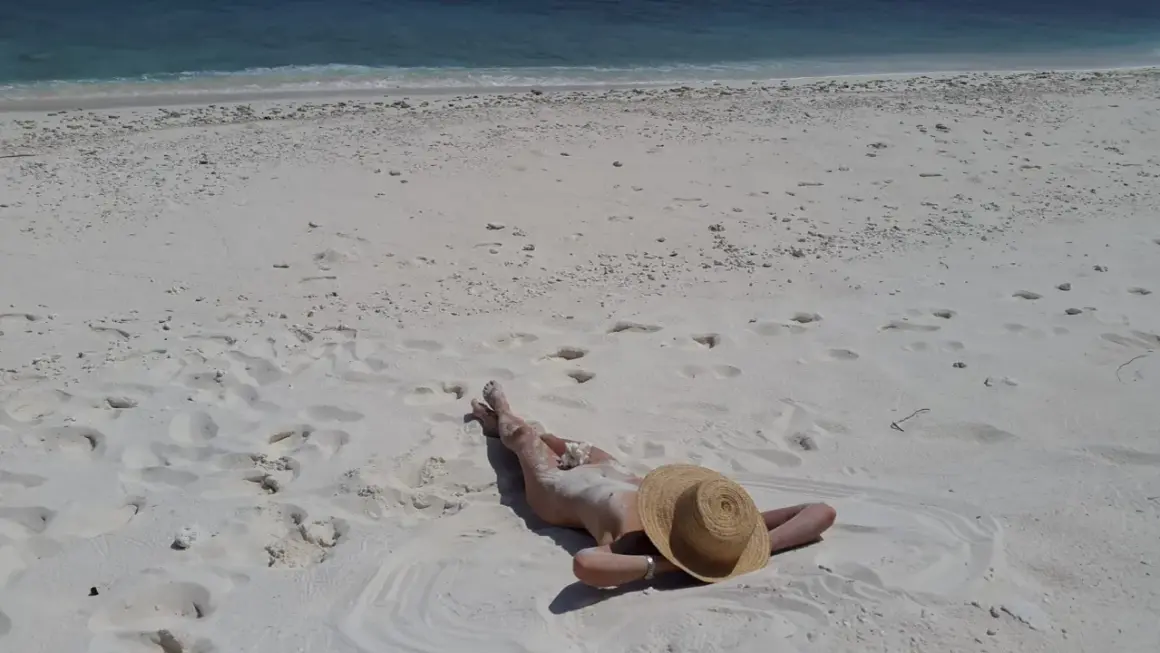 Person lying on a white sandy beach, wearing a straw hat, with the blue ocean in the background.