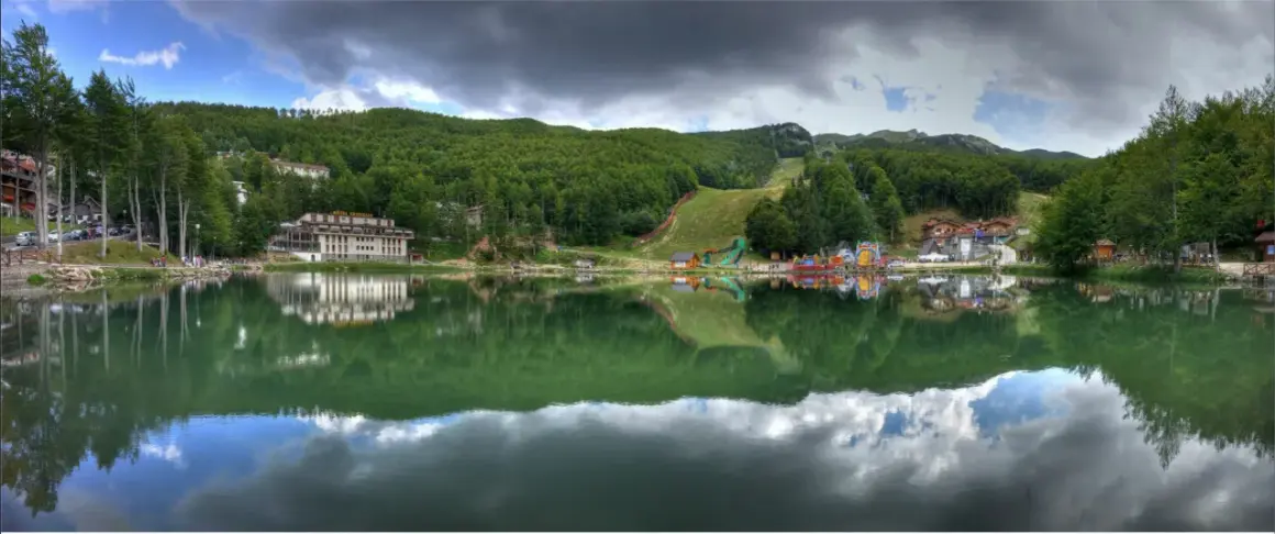 Lago alpino con riflessi di alberi e case, sullo sfondo il **passo del Cerreto** con piste da sci e giochi per bambini.