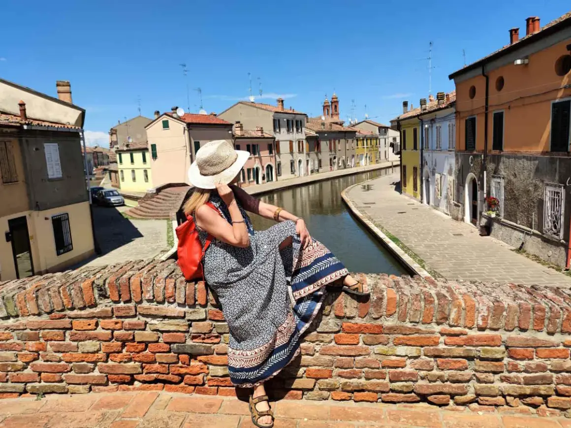 Donna con cappello di paglia e zaino rosso seduta su un ponte di mattoni a Comacchio, con canali e case colorate. Un luogo ideale per una gita con bambini.