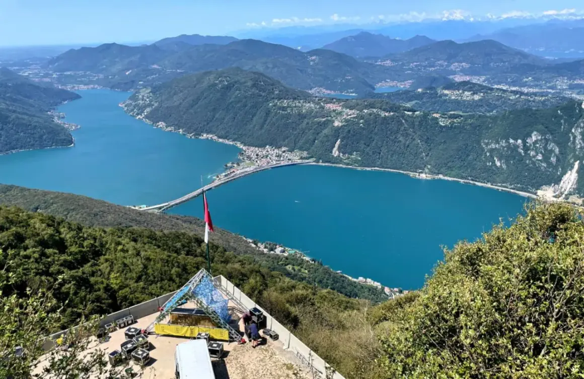 Vista panoramica del Lago di Lugano, con un ponte che lo attraversa. In primo piano, una struttura per eventi e una bandiera. Ideale per le val d'Intelvi passeggiate.