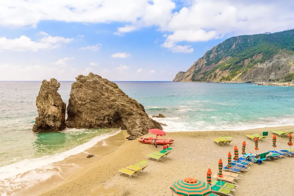 Spiaggia sabbia Liguria con scogli imponenti, mare azzurro, lettini e ombrelloni pronti per accogliere i bagnanti.