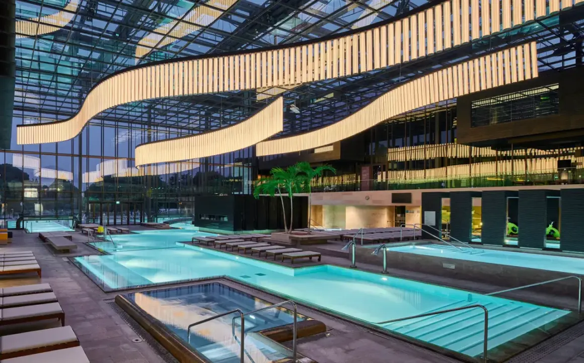 Interno moderno delle Merano Terme con piscine luminose, lettini e palme sotto un soffitto vetrato con illuminazione scenografica.