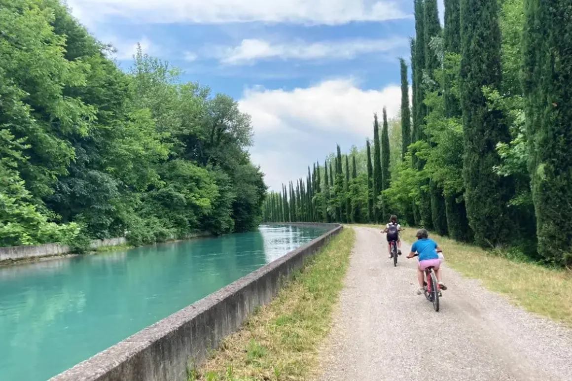 Bambini in bicicletta lungo la ciclabile Mincio, con un canale turchese e filari di cipressi.