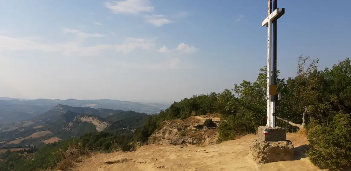 Croce in cima a un crinale roccioso, con vista panoramica sui monti dell'Emilia Romagna e un cielo azzurro con nuvole bianche.