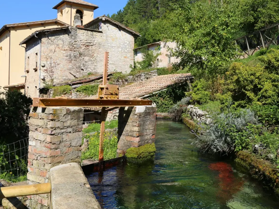 Un canale d'acqua cristallina attraversa un borgo antico, ricordando la Venezia dell'Umbria. Edifici in pietra e vegetazione rigogliosa incorniciano questo scorcio pittoresco.