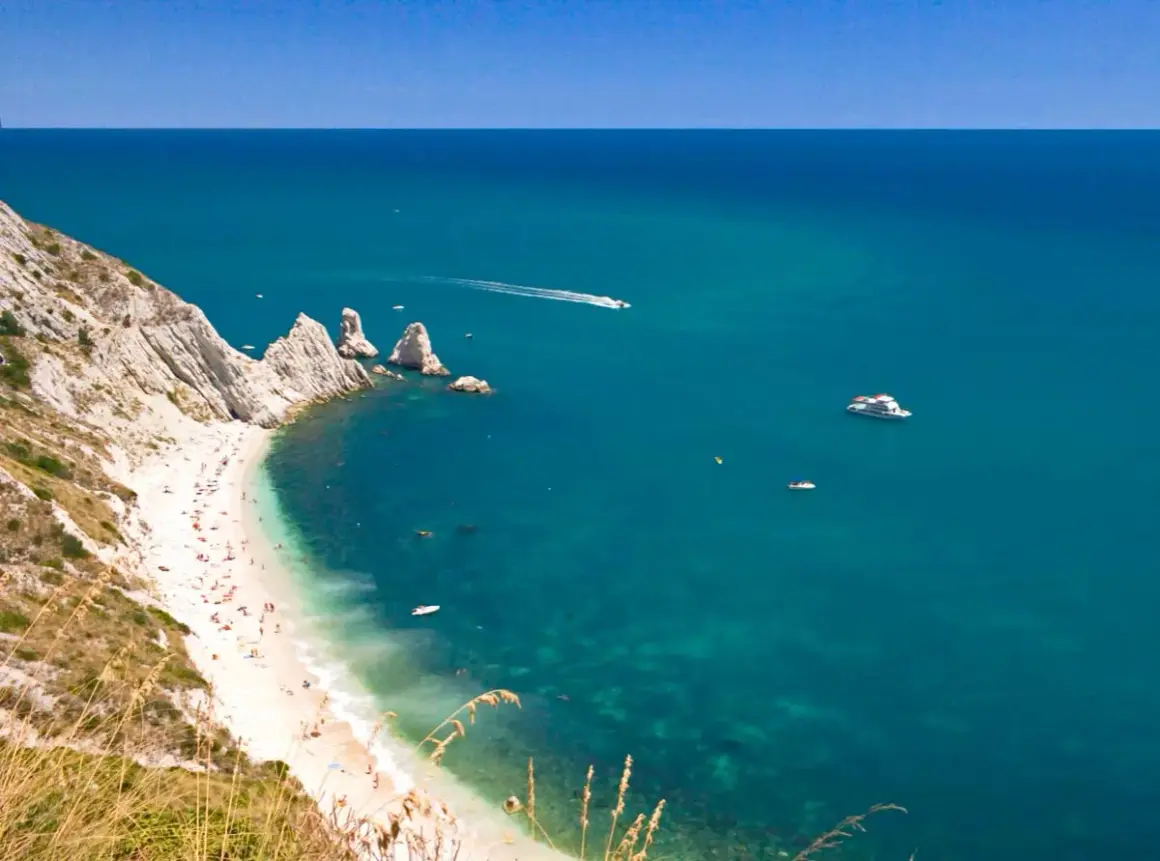 La spiaggia più bella del Conero, con sabbia bianca, mare cristallino e scogliere imponenti. Barche solcano le acque turchesi.