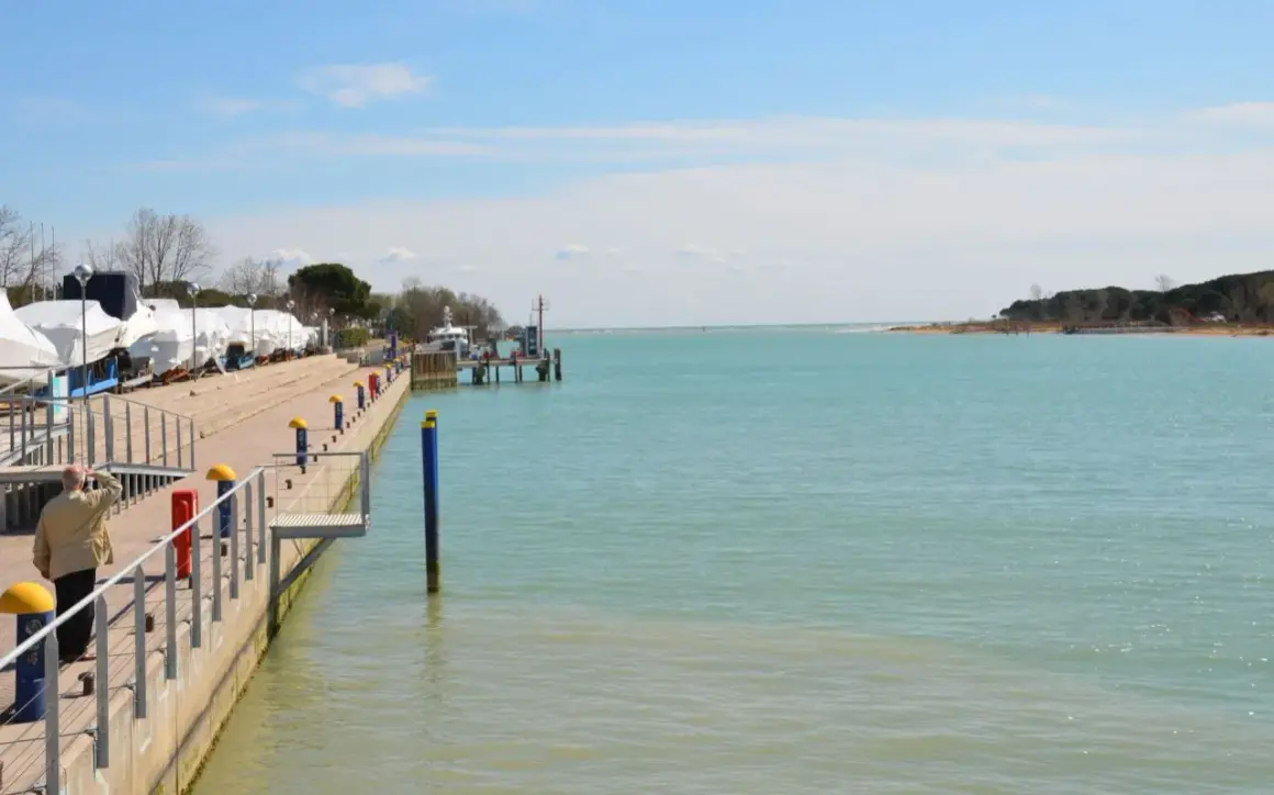 Un uomo guarda il mare calmo a Marano Lagunare, con barche coperte e un molo. La spiaggia lagunare è serena.
